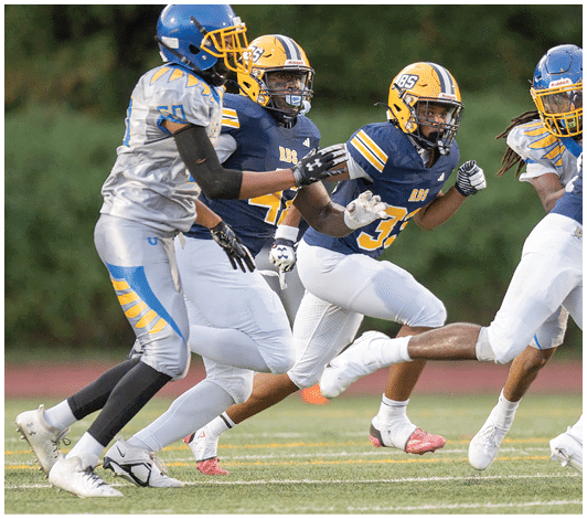A person wearing a blue and yellow football uniform with the number 57 is running on a field with other players blurred in the background. Description generated by AI