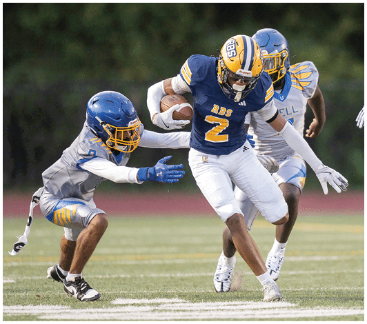 A person wearing a blue and yellow football uniform with the number 57 is running on a field with other players blurred in the background. Description generated by AI