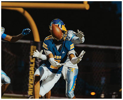 Two people in blue and yellow football uniforms are reaching for a football near a goalpost at night. Description generated by AI