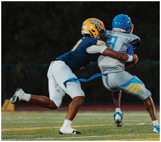 Two people wearing football helmets and uniforms are engaged in a tackle during a game with a dark background. Description generated by AI
