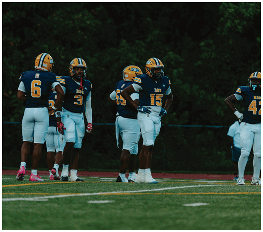 Five people wearing blue and yellow football uniforms stand on a field with a dark forest background. Description generated by AI