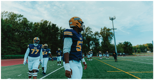 Several people wearing blue and yellow football uniforms are standing on a field with trees and a light pole in the background under a partly cloudy sky. Description generated by AI