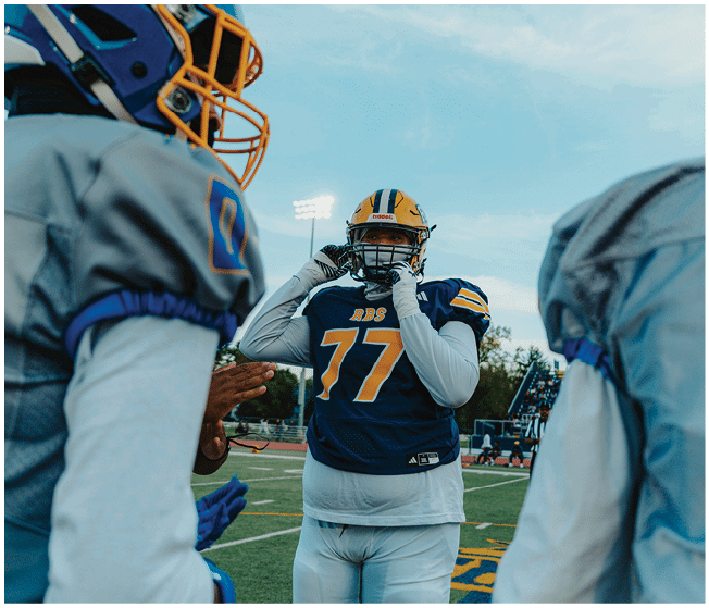 A person wearing a blue football helmet with an orange faceguard is looking towards another person in a yellow and black helmet against a clear blue sky. Description generated by AI