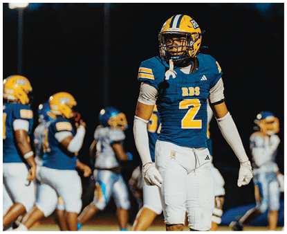 A person wearing a blue and yellow football uniform with the number 2 stands on a field at night while other players in similar uniforms are in the background. Description generated by AI