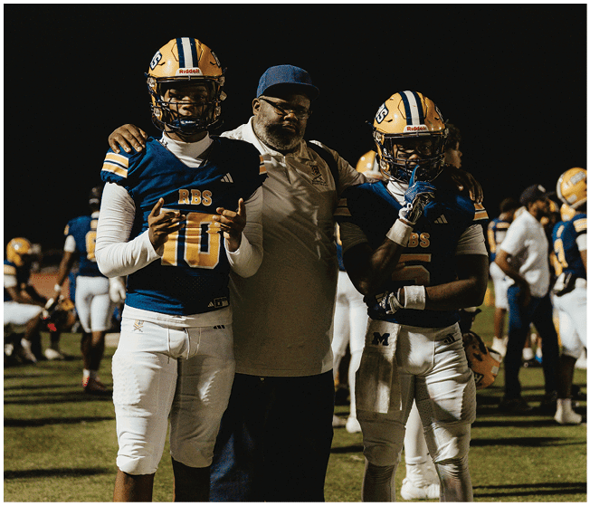 Two people in football uniforms stand on either side of a person wearing a cap and casual clothing on a football field at night. Description generated by AI