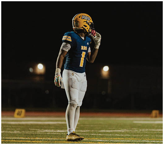 A person wearing a blue and yellow football uniform with the number one stands on a field at night, holding a hand near their mouth. Description generated by AI