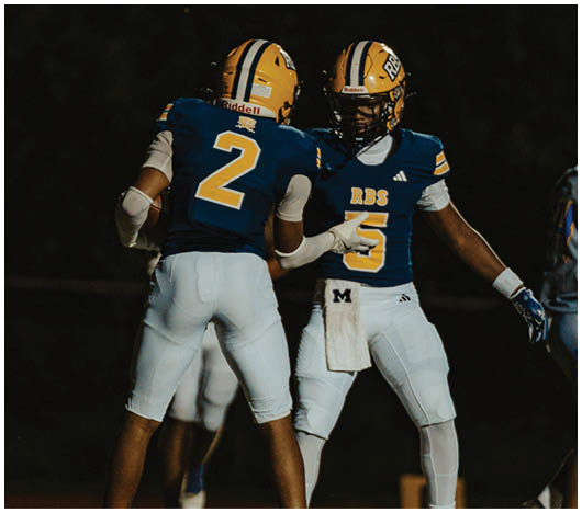 Two people in blue and white football uniforms with yellow helmets are interacting on a field while two others in gray and blue uniforms stand nearby. Description generated by AI