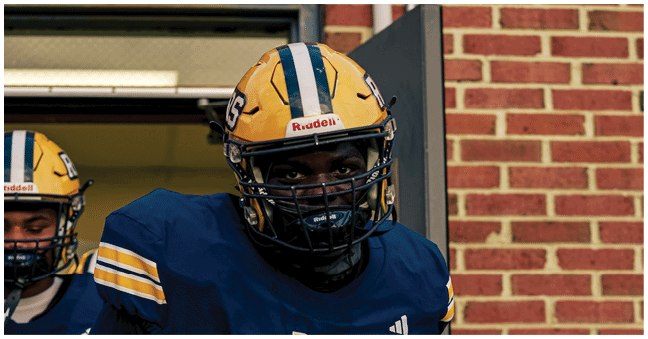 Two people wearing yellow helmets and blue sports uniforms stand in front of a brick wall and doorway. Description generated by AI
