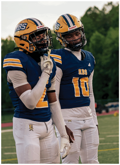Two people wearing blue and yellow football uniforms and helmets stand on a field with one person holding a hand near their face and the other looking forward. Description generated by AI