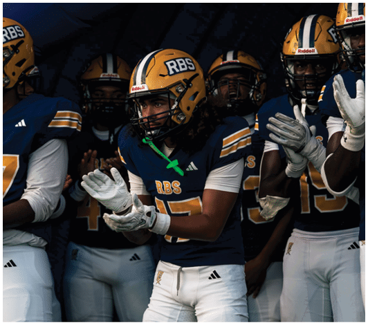 People in football uniforms and helmets are standing together, with one person in the center clapping and appearing focused. Description generated by AI
