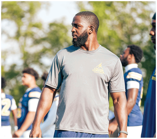 A person wearing a blue and yellow football uniform with the number 57 is running on a field with other players blurred in the background. Description generated by AI