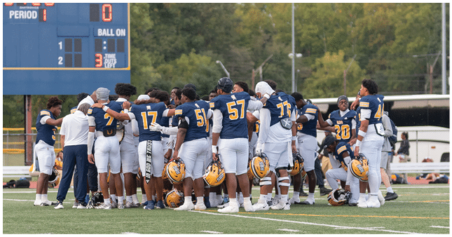 A group of football players in navy and white uniforms huddle together on a field near a scoreboard showing the first period and a score of zero. Description generated by AI