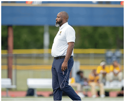 An adult with a beard wearing a white polo shirt and dark pants is walking on a sports field holding a whistle with a lanyard. Description generated by AI
