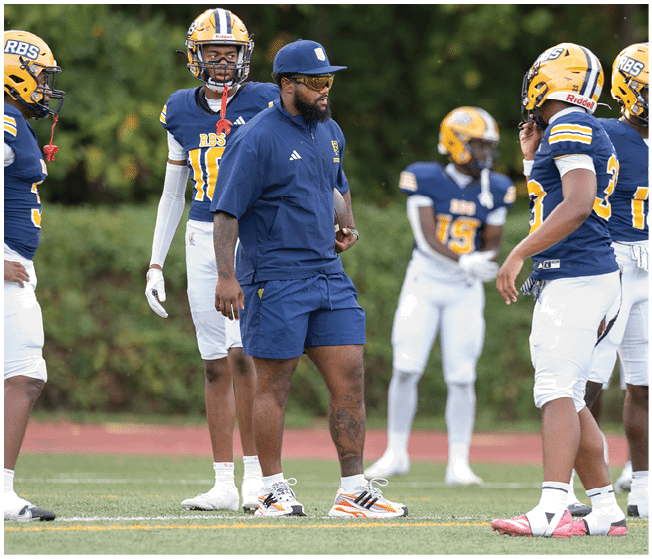 A group of people wearing blue and yellow football uniforms stand on a field with one person in a blue coach outfit giving instructions. Description generated by AI