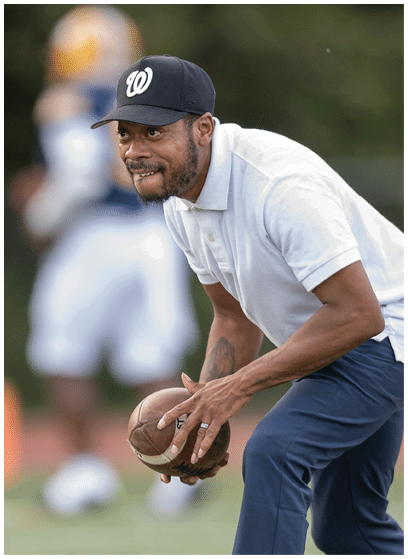 A person wearing a white polo shirt, dark pants, and a black cap is holding a football in a ready position on a sports field. Description generated by AI