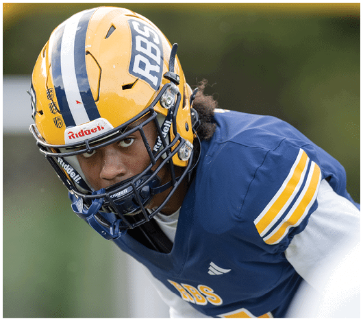 A person wearing a yellow and blue football helmet and a blue jersey with yellow and white stripes is leaning forward in an athletic stance. Description generated by AI