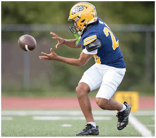 A person wearing a yellow helmet and blue jersey is reaching out to catch a football on a grassy field. Description generated by AI