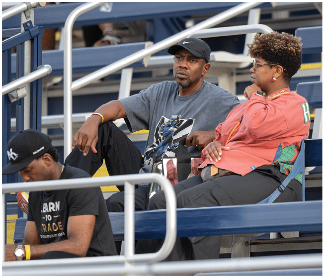 Three people sit on bleachers, with one person looking at a phone while the other two appear to be watching something off-camera. Description generated by AI
