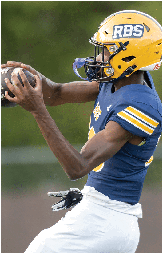 A person wearing a yellow helmet and blue sports jersey is catching a football outdoors. Description generated by AI