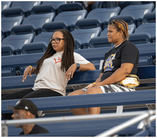 Two people with braided hair sit spaced apart on stadium bleachers, one wearing a light-colored shirt and glasses and the other wearing a dark graphic t-shirt and holding a shiny gold fanny pack. Description generated by AI