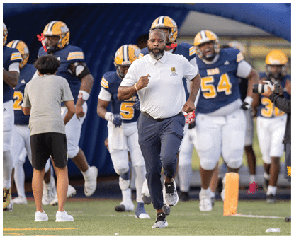 An adult in a white polo and dark pants is running on a football field with several people in football uniforms and helmets behind them. Description generated by AI