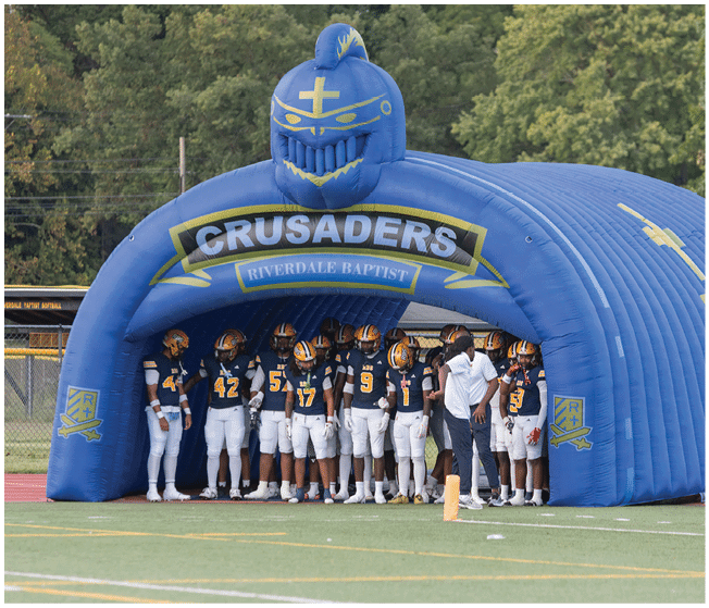 A group of people in football uniforms stand inside a large blue inflatable tunnel labeled Crusaders Riverdale Baptist on a sports field. Description generated by AI