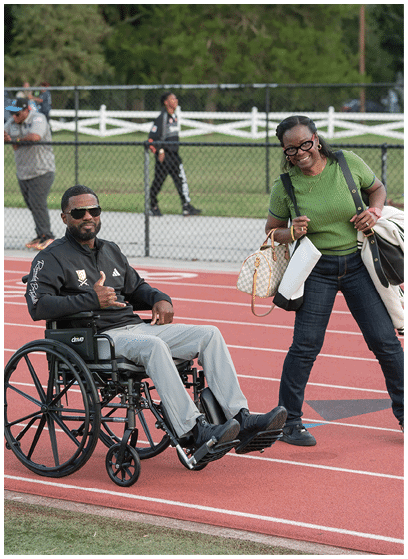 A person wearing sunglasses and a black jacket is sitting in a wheelchair on a running track while another person in casual clothing walks nearby. Description generated by AI