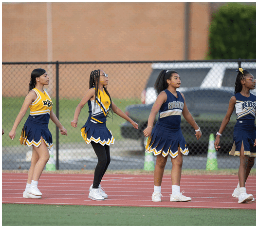 Four people in cheerleading uniforms are standing in a line on a track with a chain-link fence and parked cars in the background. Description generated by AI