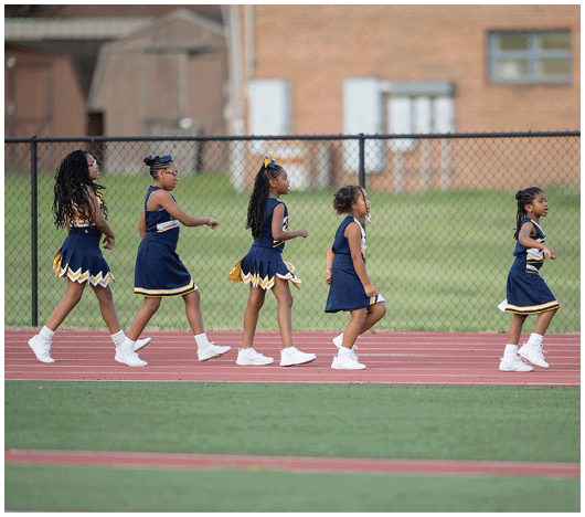 Five children in cheerleading uniforms walk in a line on a track with a chain-link fence and a building in the background. Description generated by AI