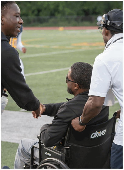 An adult wearing a white shirt and cap is pushing another adult in a wheelchair on a sports field. Description generated by AI