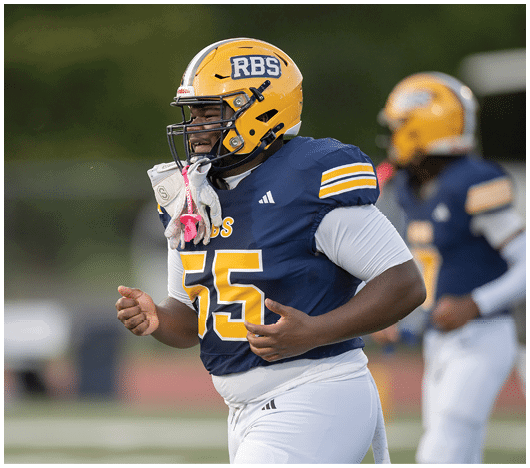 A person wearing a yellow football helmet and blue jersey with the number 55 is running on a football field with another player in the background. Description generated by AI