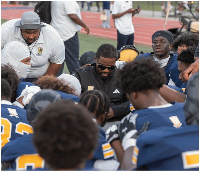 A group of people in sports uniforms and coaches are gathered closely on a field, appearing to have a focused discussion or pep talk. Description generated by AI
