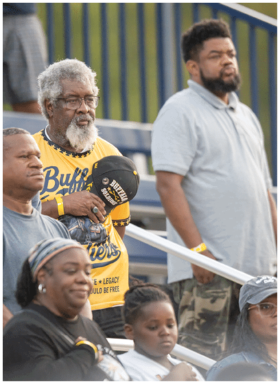 Several adults and children are seated in bleachers at an outdoor event, with one adult wearing a yellow sports jersey and holding a black cap. Description generated by AI
