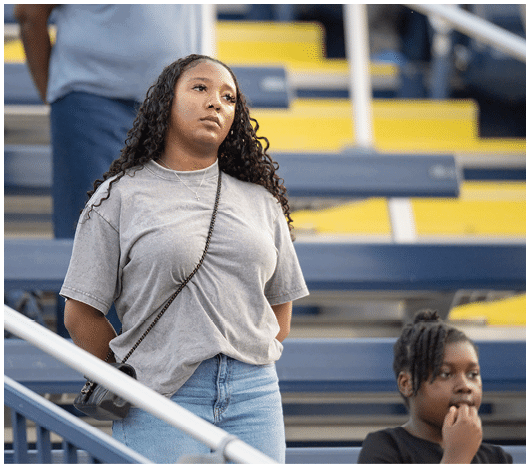 A person with long curly hair wearing a gray t-shirt and blue jeans stands with hands behind their back in front of bleachers while another person with braided hair sits nearby. Description generated by AI