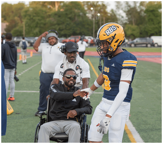 A person in a football uniform and helmet shakes hands with a person sitting in a wheelchair on a sports field with other people in the background. Description generated by AI