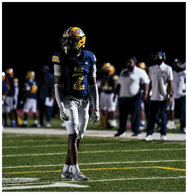 A person wearing a blue and yellow football uniform with the number 2 stands on a field at night with other people and players blurred in the background. Description generated by AI