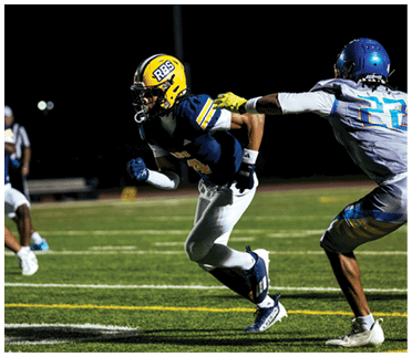 Two people wearing helmets and sports gear are engaged in an action on a dark field under a bright light. Description generated by AI
