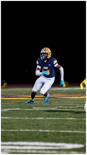 Two people in football uniforms are on a field at night with one person running toward the other. Description generated by AI