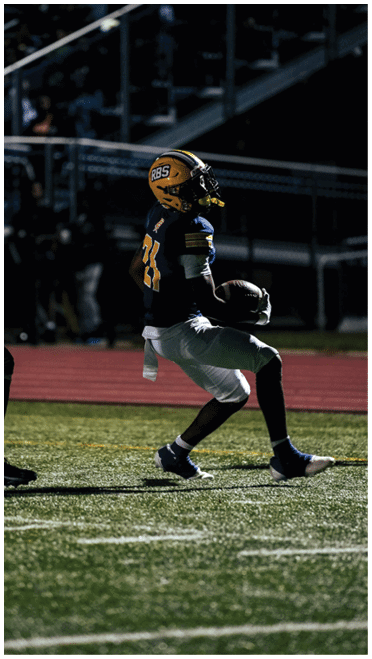 A person wearing a dark sports uniform and helmet is running on a football field while holding a football, with other players and stadium seating visible in the background. Description generated by AI