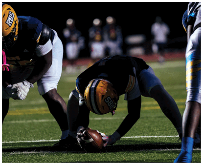 Two people in football gear are positioned on a field at night with blurred figures in the background. Description generated by AI