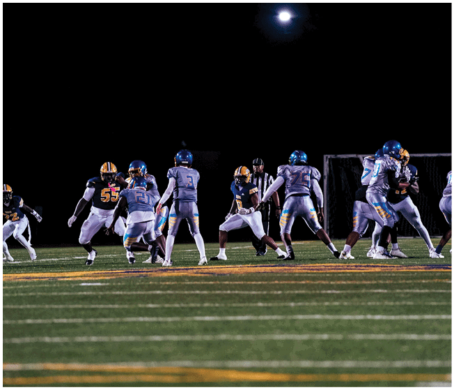 A group of people wearing football helmets and uniforms are gathered on a field under a dark night sky with a bright moon visible above. Description generated by AI