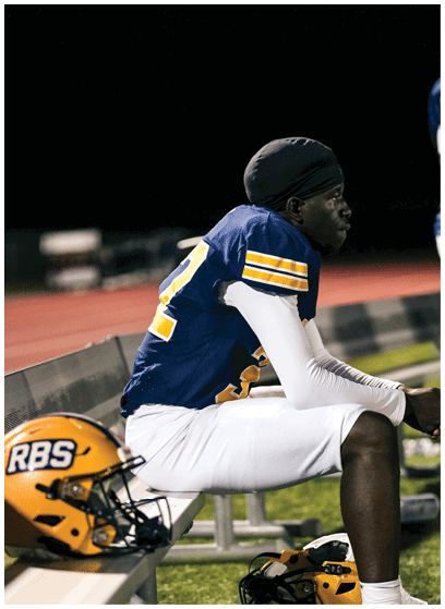 A person wearing a blue and yellow football uniform sits on a bench on a sports field with a helmet placed nearby. Description generated by AI