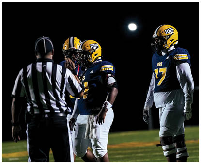 Three football players wearing yellow helmets and blue jerseys stand on a field at night with a referee in the foreground and a bright light in the background. Description generated by AI