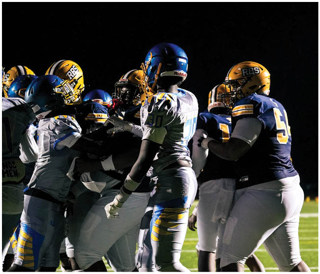 A group of people wearing football helmets and uniforms are gathered closely together against a dark background. Description generated by AI