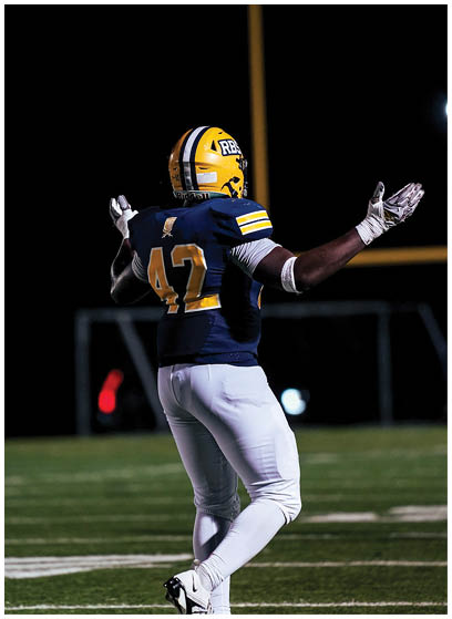 A person wearing a yellow helmet and blue football jersey with the number 42 is standing on a football field at night with arms raised. Description generated by AI