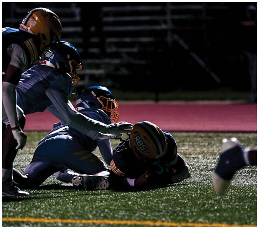 Three people wearing football helmets and uniforms are crouched in a ready position on a field at night with empty bleachers in the background. Description generated by AI