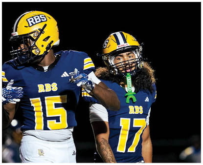 Two people wearing blue and yellow football uniforms with helmets are walking on a field at night with blurred figures in the background. Description generated by AI