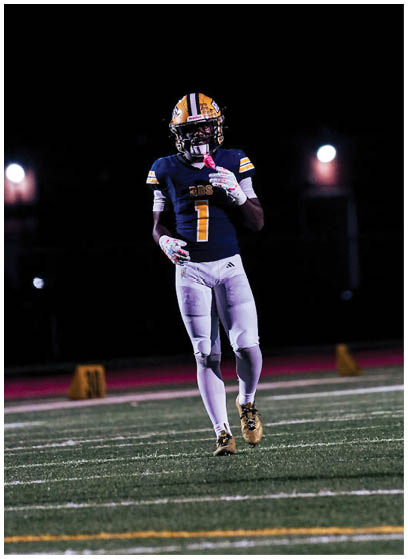 A person wearing a football uniform with a helmet and gloves is standing on a lit football field at night. Description generated by AI