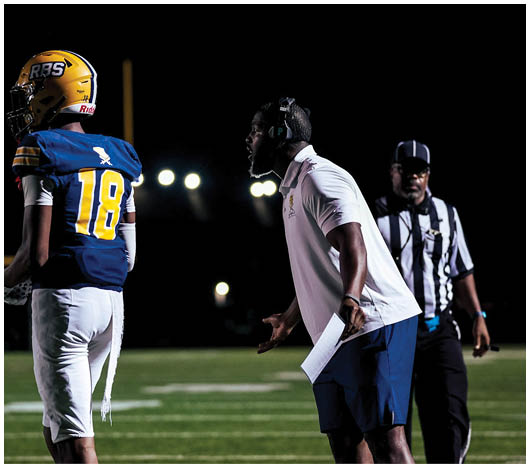 A person wearing a headset and holding a paper is shouting towards a player in a football uniform with the number 18, while a referee stands nearby on a lit football field at night. Description generated by AI