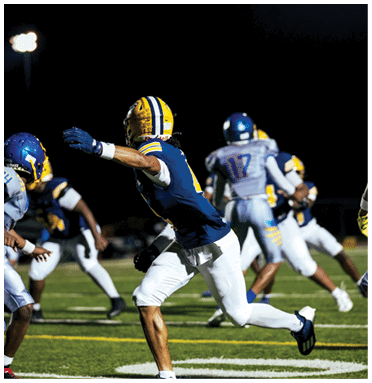 People wearing football uniforms are actively playing on a well-lit field at night. Description generated by AI